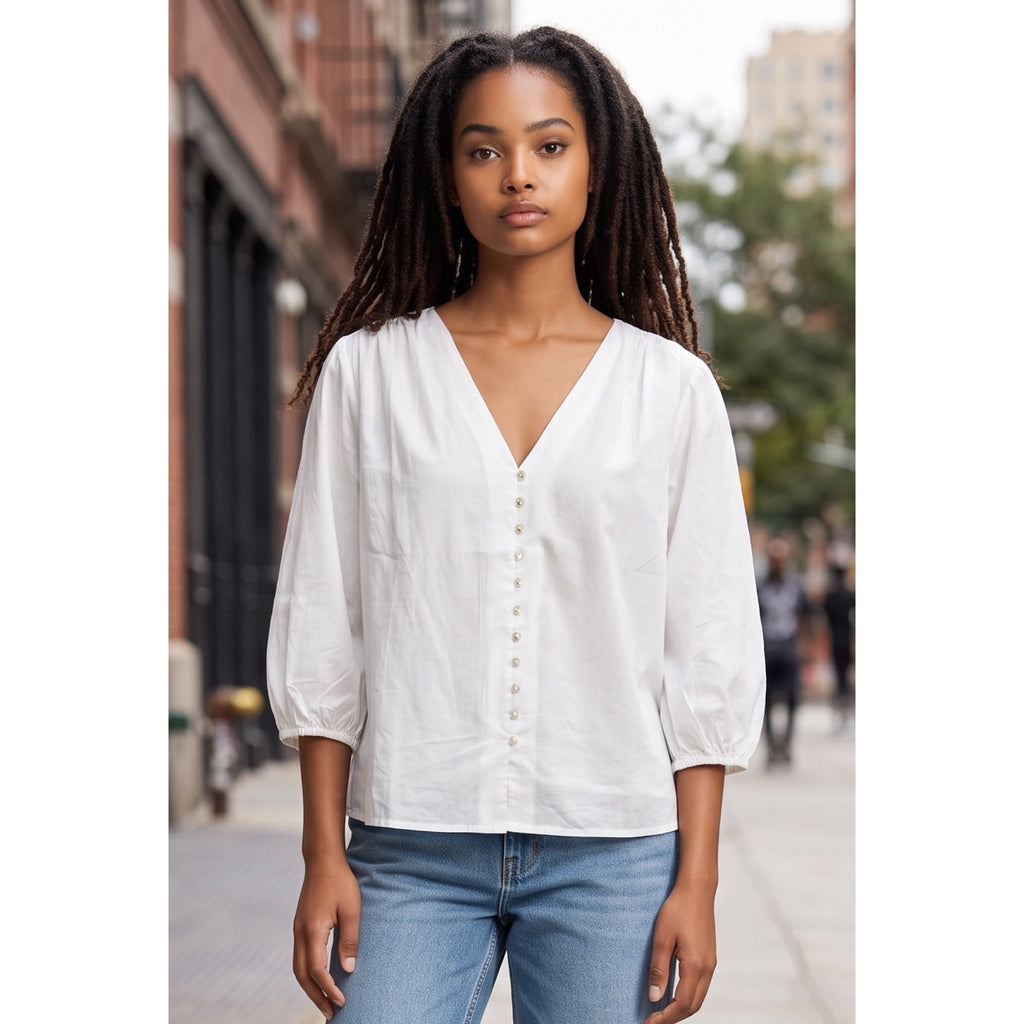 Vista cercana de blusa blanca con escote en pico, botones delanteros, frunces en los hombros y mangas abullonadas.