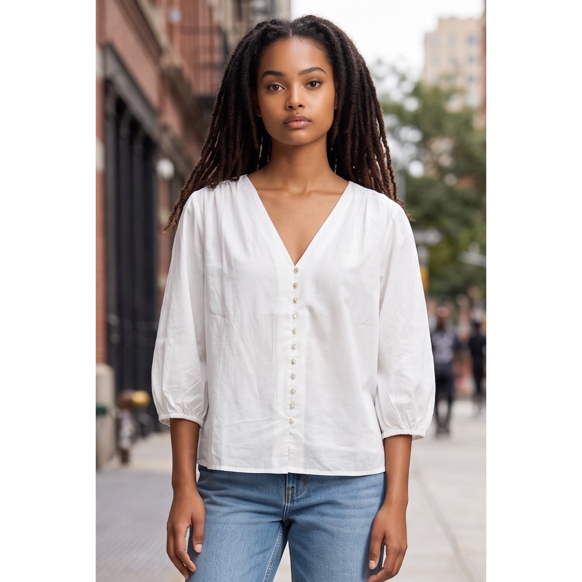 Vista cercana de blusa blanca con escote en pico, botones delanteros, frunces en los hombros y mangas abullonadas.