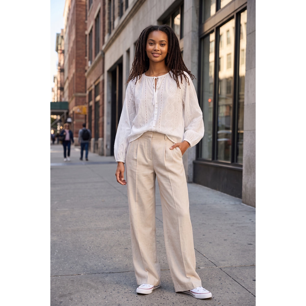 Modelo con blusa blanca de bordado inglés y lazada, llevada parcialmente por dentro de un pantalón beige.