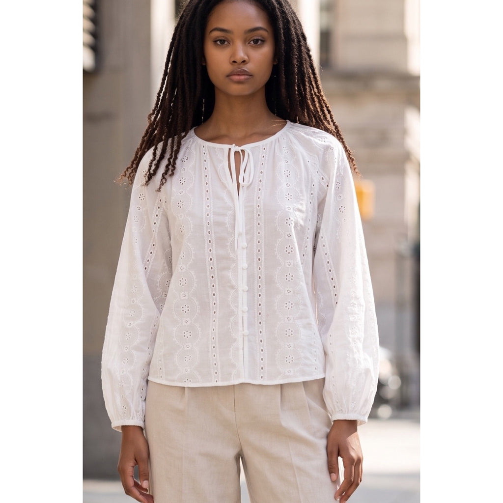 Vista cercana de la blusa blanca con lazada en el cuello, botones delanteros y mangas largas bordadas.