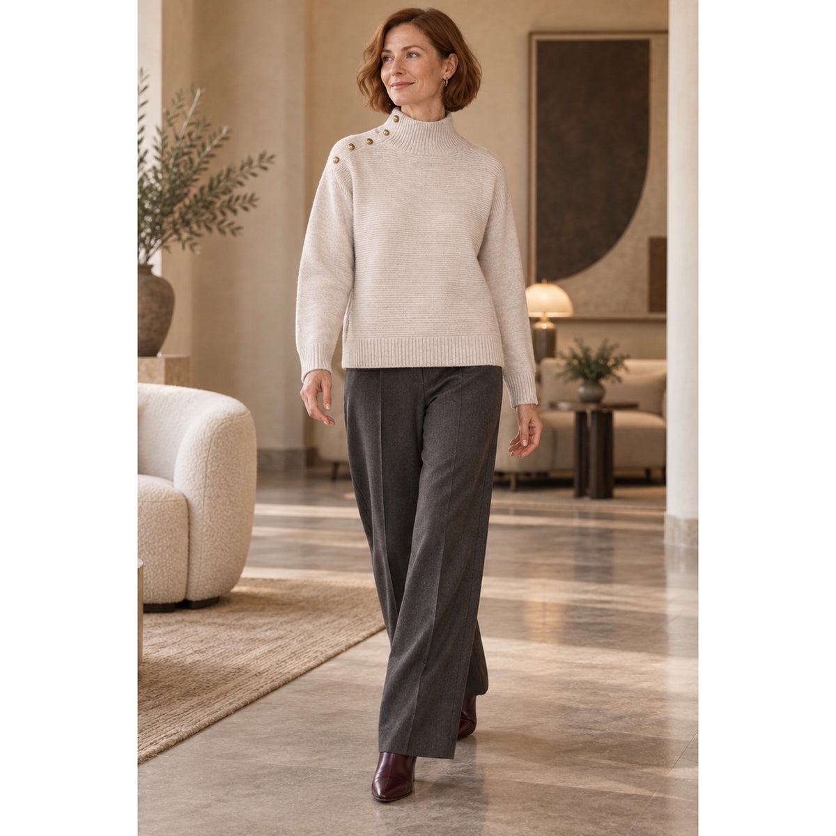 Modelo con jersey beige de cuello alto, botones dorados en el lateral del cuello y el hombro, y pantalón gris.